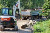 a digger and truck driving down a dirt road next to a forest