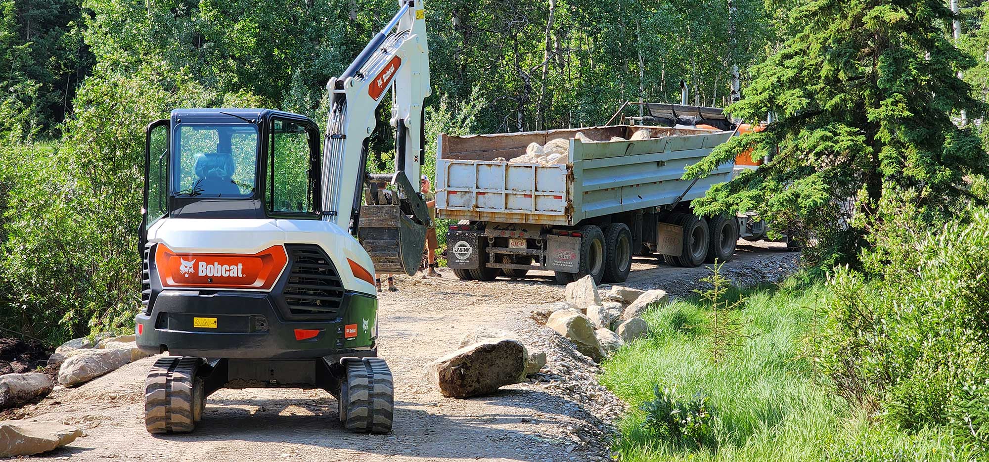 a digger and truck driving down a dirt road next to a forest